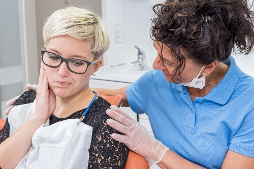 A patient rubs her cheek in pain while a dental hygienist stands next to her for comfort.
